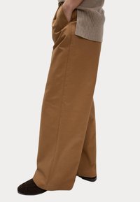 Breda bruna byxor i ett mjukt tyg. Har sidfickor och en avslappnad passform, ihop med mörka texturerade tofflor.