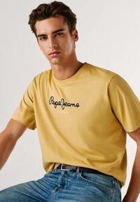 Camiseta de algodón amarilla con mangas cortas, que presenta el logo "Pepe Jeans London" en negro. Llevada con jeans de mezclilla azul claro y un cinturón negro.