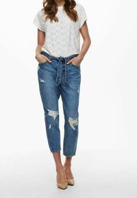 Top blanco ligero con diseño de ojetes, combinado con jeans azul desgastados de corte holgado y tacones beige. Detalle de cintura anudada en los jeans.