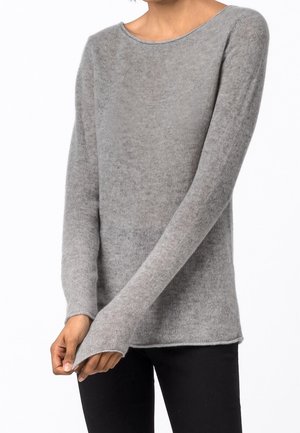 Nainen, joka käyttää vaaleanharmaata, pitkähihnaista neulepaitaa leveällä kaula-aukolla ja mustia housuja, seisoo valkoista taustaa vasten.