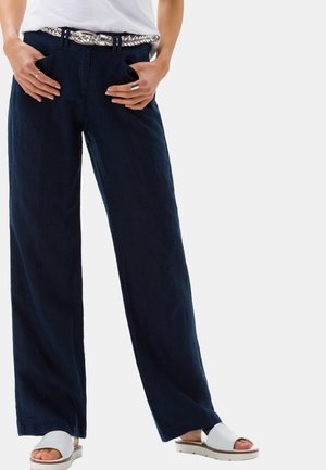 Persoon draagt marineblauwe wijde broek met een zilveren gevlochten riem, witte open-toe slide sandalen en houdt handen in de zakken.