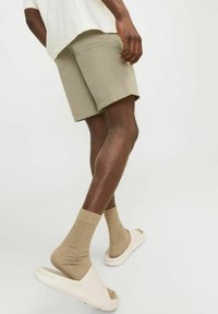 Beige teksturēti šorti ar brīvu piegulšanu, beige potīšu zeķes un baltas slaidas sandalēs, skatīts no sāniem. Ikdienas apģērba dizains.