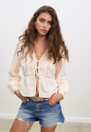 Cremefarbendes Blusenshirt mit V-Ausschnitt und vordere Schleife, verziert mit gestickten Details und elastischen Ärmeln, kombiniert mit blauen Jeansshorts.