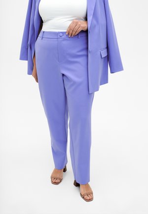 Donna che indossa pantaloni sartoriali lavanda e un blazer abbinato su una maglietta bianca, abbinati a sandali con tacco marroni su sfondo bianco.