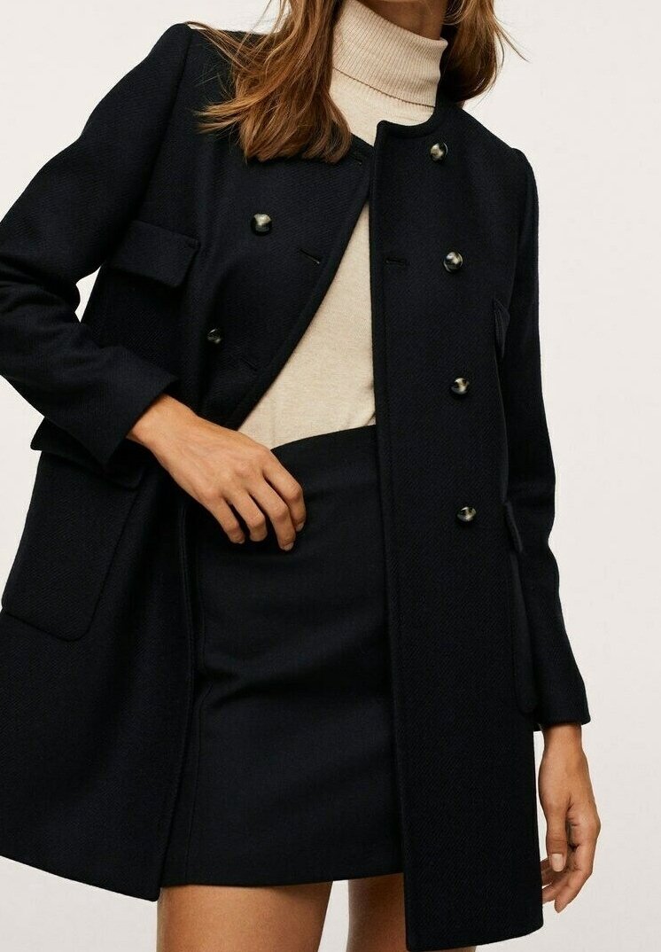 Femme portant un manteau noir croisé sur un col roulé beige et une jupe noire, une main reposant sur sa taille.