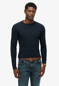 Marineblaue Langarm-Henley-Bluse aus Baumwolle, mit einer Knopfleiste aus drei Knöpfen und einem dezenten Logo auf der Brust, kombiniert mit Jeans.