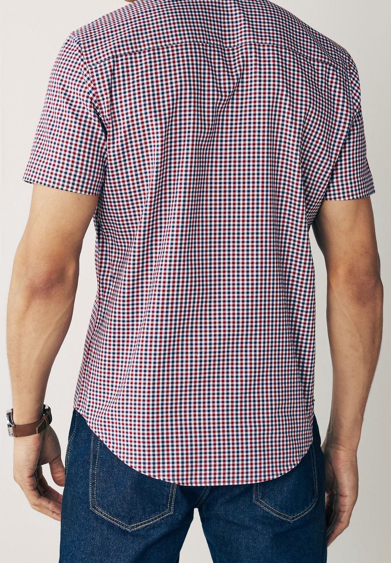 Homme portant une chemise à manches courtes à carreaux rouges, blancs et bleus et un jean bleu, vu de dos avec une montre-bracelet au poignet gauche.