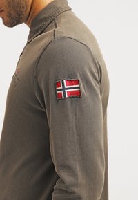Un homme portant une chemise à manches longues marron avec un patch du drapeau norvégien cousu sur le bras supérieur, vu de l'épaule au cou.