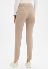 Beige slim-fit broek met een gladde textuur, twee achterzakken en gestikte details; gecombineerd met witte sneakers.