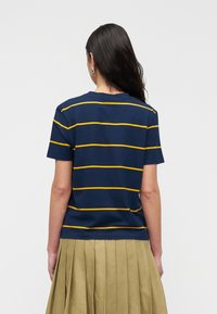 Μπλε ναυτικό T-shirt με οριζόντιες κίτρινες ρίγες, κατασκευασμένο από βαμβάκι, με κοντά μανίκια και στρογγυλή λαιμόκοψη, φορεμένο με πλισέ μπεζ φούστα.