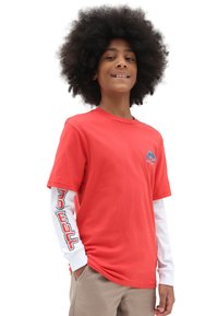 T-shirt di cotone rosso con maniche corte indossata sopra una camicia a maniche lunghe bianca, con testo grafico nero e rosso sulle maniche.