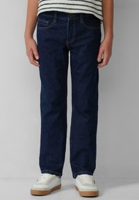 Jean en denim bleu foncé avec une coupe droite, poches frontales et détails de couture visibles, associés à des baskets blanches.