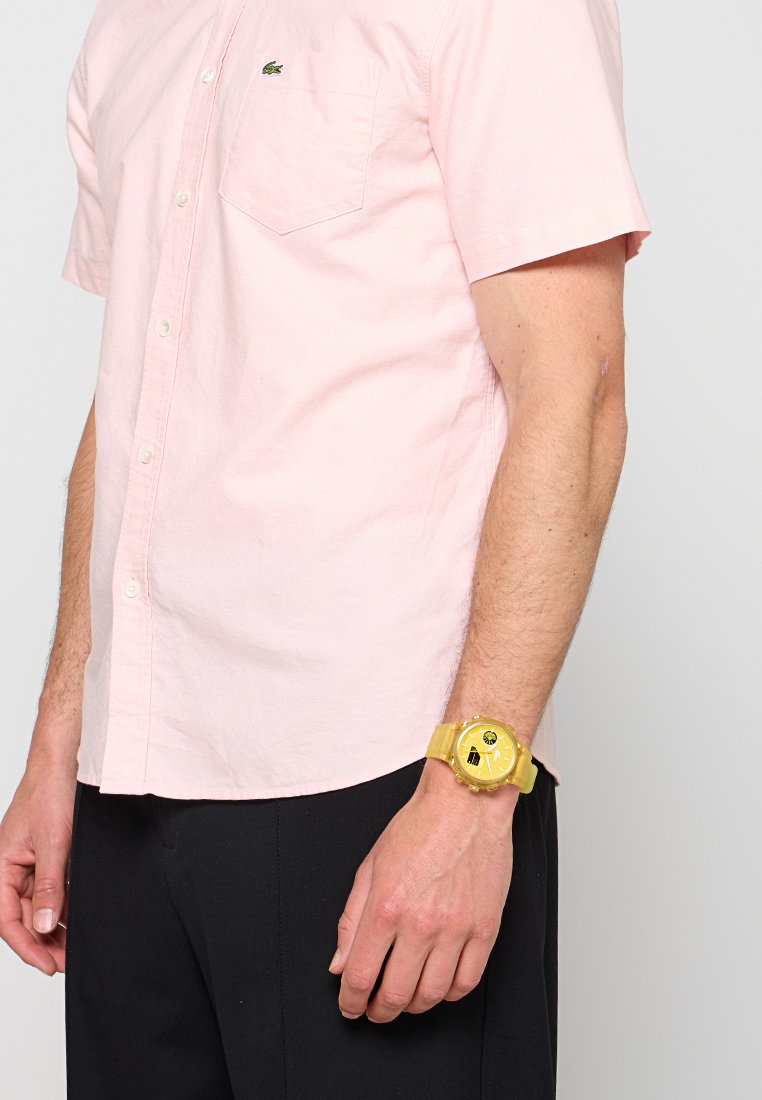 Hombre con camisa rosa claro de manga corta con botones, pantalones negros y un gran reloj de pulsera amarillo. Solo son visibles el torso y parte de un brazo.