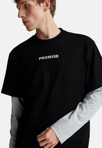 Mladý muž nosí černé tričko s bílým nápisem "PROMISE" přes šedou dlouhou rukávovou košili a dívá se vlevo na bílém pozadí.