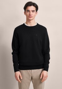 Junger Mann steht, trägt einen schwarzen Pullover über einem weißen Hemd und beigen Hosen vor einem schlichten beigen Hintergrund.