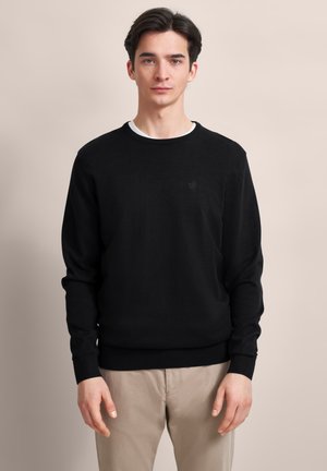  jeune homme debout, portant un pull noir sur une chemise blanche et un pantalon beige, devant un fond beige uni.