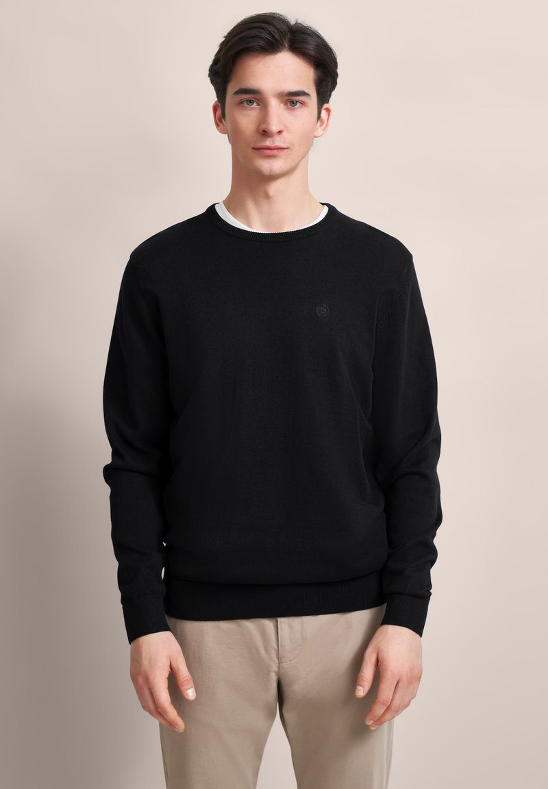 Junger Mann steht, trägt einen schwarzen Pullover über einem weißen Hemd und beigen Hosen vor einem schlichten beigen Hintergrund.