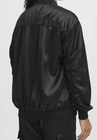 Schwarze Satin-Bomberjacke mit gerafftem Rücken und Ärmeln, gerippten Bündchen und glatter Textur. Verfügt über eine lockere Passform und ein minimalistisches Design.