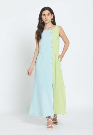 Žena stojí oblečená v bezrukávových šatách dĺžky po zem, s ľavou stranou svetlomodrou a pravou stranou svetlozelenou, s rovnako farebnými gombíkmi vpredu.