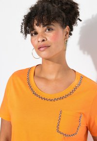 Orange bomulds t-shirt med rund halsudskæring dekoreret med rhinsten og en lomme med et lignende design.