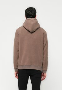 Man wearing a brown fleece hoodie with the hood down, standing facing a white wall, hands relaxed by sides, black pants visible.