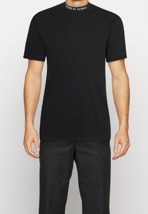 Homme portant un t-shirt noir uni à manches courtes avec un petit texte blanc sur le col et un pantalon noir.