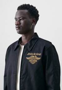 Black Dickies jacket featuring golden embroidered logo with wings, collar, and a zipper closure. Worn over a light-colored shirt.