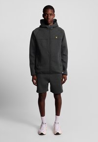 Felpa con zip grigia scura abbinata a pantaloni corti, realizzata in un materiale morbido. Presenta un logo giallo a contrasto sul petto e due tasche laterali.