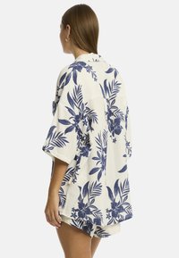 Una donna è in piedi con le spalle alla macchina fotografica, indossando una camicia a maniche corte con un motivo floreale e pantaloni corti coordinati.