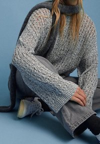 Personne assise par terre portant un pull gris ample en maille, un pantalon gris, des chaussettes foncées, des baskets blanches et une écharpe sombre sur un fond bleu.