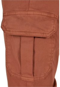 Poche cargo en tissu couleur rouille avec rabat et coutures visibles sur un pantalon.