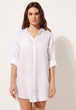 Ung kvinde med langt brunt hår iført hvid oversized hørskjortekjole og gyldne øreringe, stående mod en ensfarvet lys baggrund.