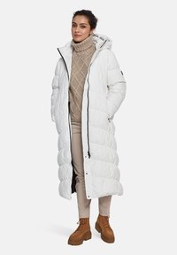 Lang witte pufferjas met een capuchon, met een gequilt ontwerp, zwarte rits en zijzakken, gedragen over een beige kabelgebreide trui en beige broek.
