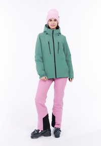 Chaqueta impermeable verde con cremalleras y acentos negros, combinada con pantalones de esquí rosas que tienen secciones inferiores negras y botas de esquí negras.