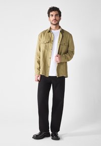 Camisa verde oliva de botones con dos bolsillos en el pecho, superpuesta sobre una camiseta blanca, combinada con pantalones negros de pierna recta y zapatos negros.