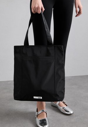 Borsa tote nera in nylon con una forma piatta e dettagli testurizzati. Presenta una tasca frontale e un logo sul manico. Scarpe argento sotto.