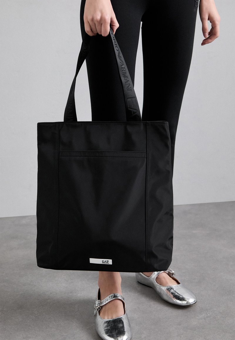 Borsa tote nera in nylon con una forma piatta e dettagli testurizzati. Presenta una tasca frontale e un logo sul manico. Scarpe argento sotto.