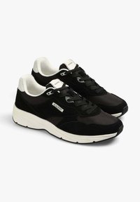 Sneakers neri che presentano una combinazione di materiali lisci e testurizzati, dettagli bianchi, stringhe e una suola spessa bianca. Etichetta del marchio visibile.