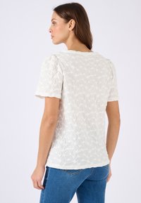 Witte textuur blouse met korte mouwen, voorzien van een bloemenpatroon en licht gepofte schouders, gecombineerd met blauwe spijkerbroek.