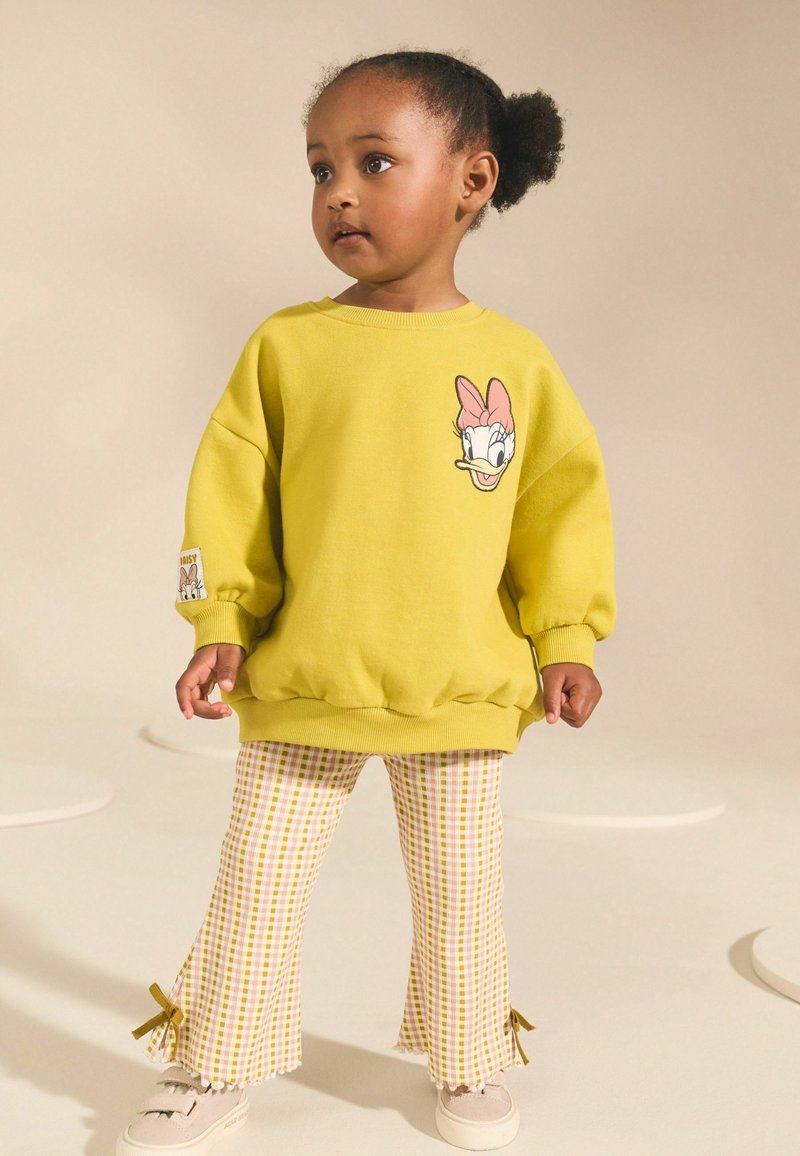 Tout-petit portant un sweat-shirt jaune avec un visage de canard de dessin animé et un pantalon à carreaux avec de petits nœuds aux chevilles, debout sur un fond clair.