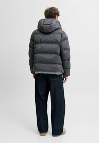 Szara kurtka puchowa z dużym kapturem, pikowanymi sekcjami i błyszczącym wykończeniem, w połączeniu z ciemnymi, szerokimi dżinsami. Minimalistyczny design.