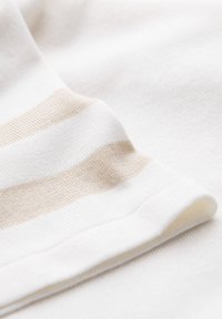 Blødt hvidt stof med beige vandrette striber, close-up viser glat tekstur og syet søm, sandsynligvis en del af et klædningsstykke.