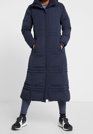 Person steht mit den Händen in den Taschen, trägt einen langen marineblauen Steppmantel, marineblaue Leggings und schwarze und weiße Turnschuhe vor einem schlichten Hintergrund.