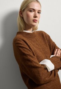 Pull en maille marron avec un col montant à côtes et des détails côtelés sur les côtés. Superposé sur une chemise blanche à col, avec des poignets retroussés.