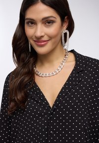 Blusa nera con pois bianchi, con scollatura a V, abbinata a una collana di catena argentata spessa e grandi orecchini a cerchio con strass.