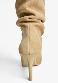 Beige velúr stiletto bokacsizma laza kialakítással, magas sarokkal és texturált felülettel, sima bézs talppal.