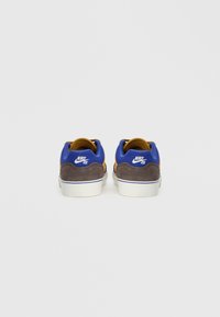 Adidași din velur albastru și maro cu o talpă albă. Prezintă un logo vizibil pe călcâi și texturi contrastante între materiale.