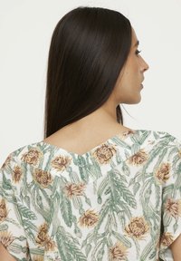 Chemisier à motifs floraux avec un col en V, présentant principalement des fleurs vertes et jaunes sur un fond blanc. Tissu doux avec des manches courtes.