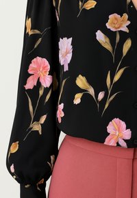 Blusa nera con maniche lunghe caratterizzata da un motivo floreale in tonalità di rosa e giallo. Il tessuto ha una texture liscia e una chiusura a bottoni.