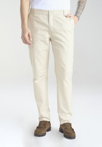 Beżowe, slim-fit spodnie z mieszanki bawełny, z pojedynczym zapięciem na guzik, kieszeniami z przodu i widocznymi detalami przeszyć, w połączeniu z brązowymi butami.